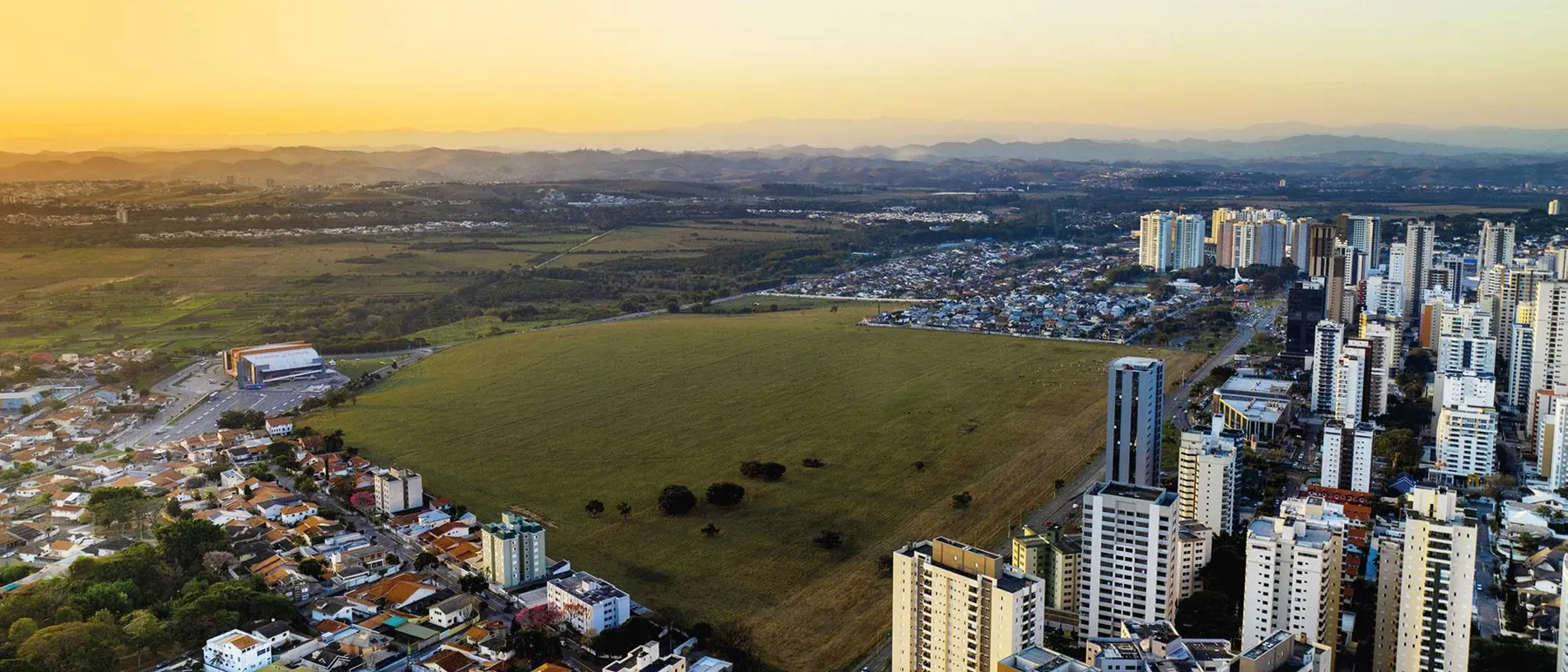 Foto aérea ao entardecer: o terreno gramado do Parque Una ocupa o centro do quadro, cercado pelas torres do Jardim Aquarius à direita e por bairros residenciais à esquerda, com a Serra da Mantiqueira no horizonte.