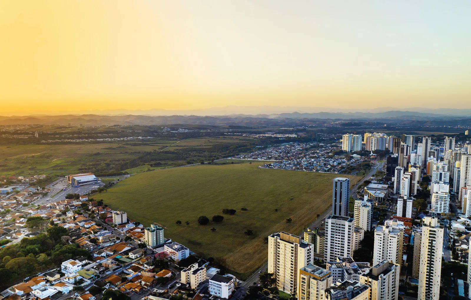 Vista aérea do terreno do Parque Una ao entardecer: uma gleba verde vazia cercada de ruas, com as torres do Jardim Aquarius de um lado, casas térreas do outro e a Serra da Mantiqueira no horizonte.